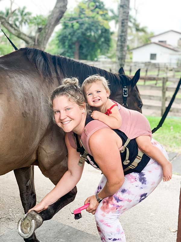 Mother smiling and picking out horse hoof with toddler on her back